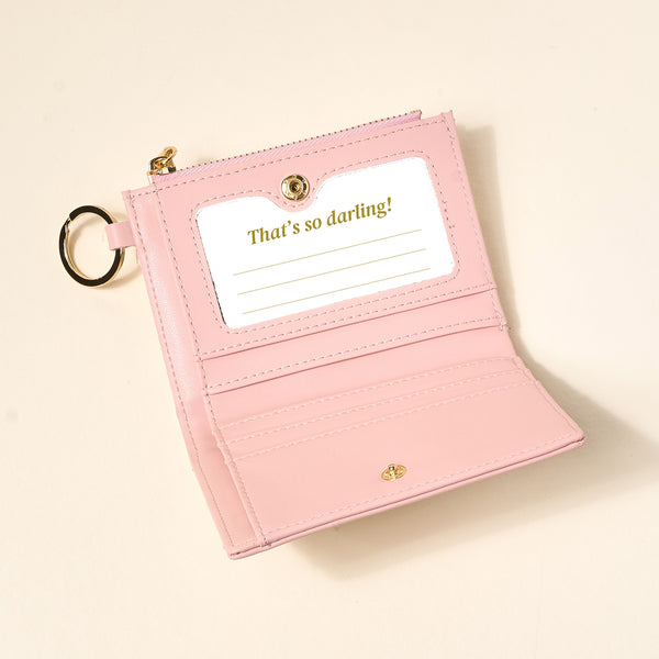 The Darling Effect’s Everbloom Dusty Rose Luxe Pocket Wallet, crafted from vegan leather, features a gold key ring, card slots, and an ID window with “That’s so darling!” in gold text—all shown on a light neutral background.
