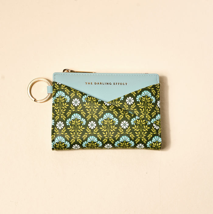 The Everbloom Forest Green Luxe Pocket Wallet by The Darling Effect is a compact vegan leather pouch with a criss-cross front, blue flap labeled with the brand, keyring, and green, blue, and white floral patterns on dark.