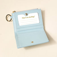 The Everbloom Forest Green Luxe Pocket Wallet by The Darling Effect is open, displaying card slots and a clear ID window with "Thats so darling!" text. A gold keychain ring is attached to the side.