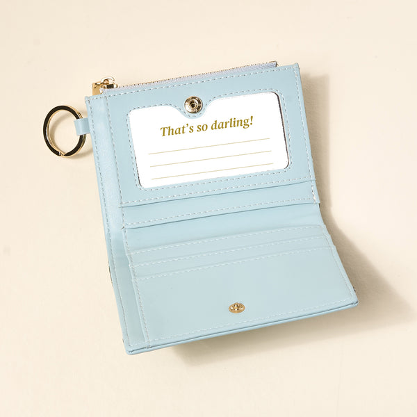 The Everbloom Forest Green Luxe Pocket Wallet by The Darling Effect is open, displaying card slots and a clear ID window with "Thats so darling!" text. A gold keychain ring is attached to the side.