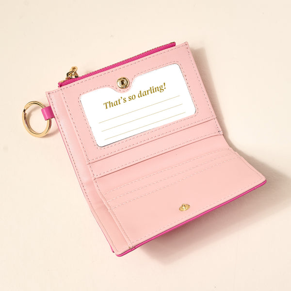 The Darling Effect’s Hot Pink Luxe Pocket Wallet features vegan leather, a gold keychain, zipper detail, card slots, a clear ID window, and “Thats so darling!” text—making it the perfect everyday accessory.