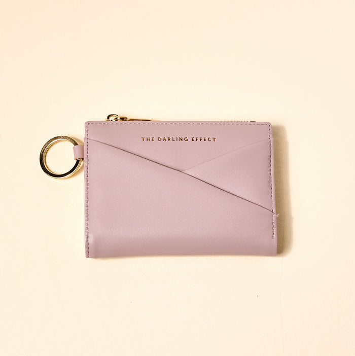 The Darling Effect's Lavender Luxe Pocket Wallet is a pastel lavender vegan leather wallet with a gold keyring, zip, and “THE DARLING EFFECT” embossed in gold on the front, shown on a light cream background.