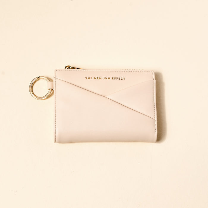 The Darling Effect Oat Luxe Pocket Wallet is a compact vegan leather wallet in light beige, featuring a gold zipper, key ring, and "THE DARLING EFFECT" in gold on the front, displayed on a plain light background.