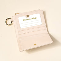 The Darling Effect's Oat Luxe Pocket Wallet, in pale pink vegan leather, features a criss-cross design, snap closure, multiple card slots, a clear ID window with “That’s so darling!” in gold script, and an attached metal key ring.