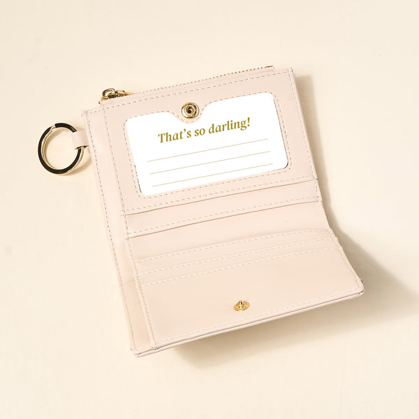 The Darling Effect's Oat Luxe Pocket Wallet, in pale pink vegan leather, features a criss-cross design, snap closure, multiple card slots, a clear ID window with “That’s so darling!” in gold script, and an attached metal key ring.