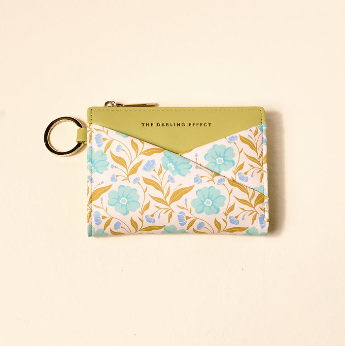 The Darling Effect Poppin' Petals Aqua Luxe Pocket Wallet: a compact vegan leather wallet with gold keyring, green flap, and floral criss-cross design in blue, yellow, and white. "THE DARLING EFFECT" is printed in gold on the green section.