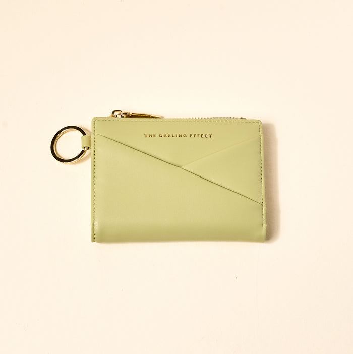 The Sage Luxe Pocket Wallet by The Darling Effect features light green vegan leather, a gold zipper, key ring, and gold-embossed branding—stylish and functional for everyday use, shown here on a beige background.