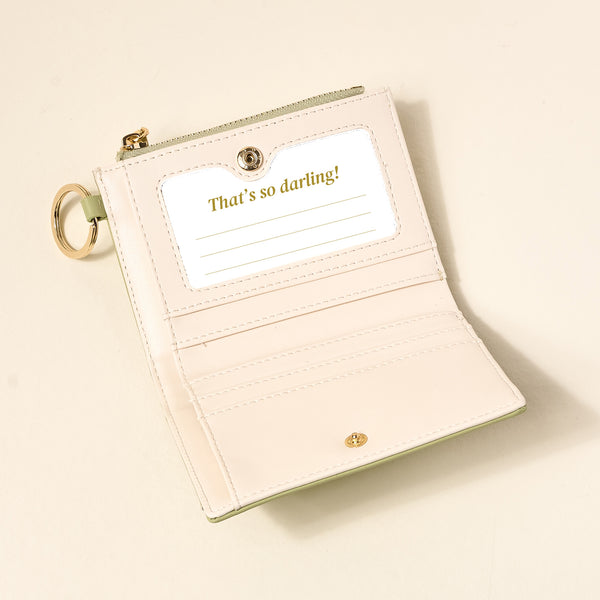 The Darling Effect's Sage Luxe Pocket Wallet is a light pink vegan leather wallet with pale green trim and a keychain. It has a clear ID window with "Thats so darling!", multiple card slots, and is perfect for everyday organization.