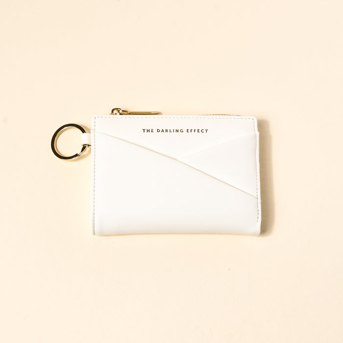 The Darling Effect White Luxe Pocket Wallet, made from vegan leather, features a gold zipper, key ring, and gold-printed logo on the front, shown on a light beige background.