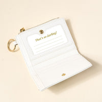 The Darling Effect’s White Luxe Pocket Wallet, a small vegan leather accessory with a gold keyring, sits on a beige surface. Open, it reveals an interior pocket and card slot stamped “That’s so darling!” in gold.