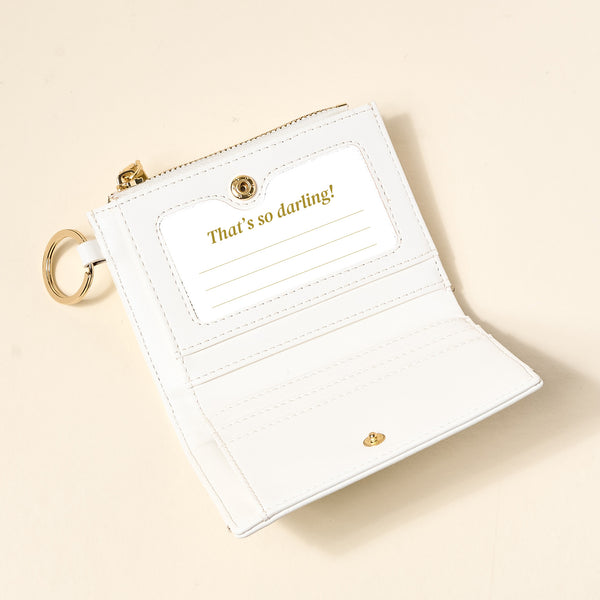 The Darling Effect’s White Luxe Pocket Wallet, a small vegan leather accessory with a gold keyring, sits on a beige surface. Open, it reveals an interior pocket and card slot stamped “That’s so darling!” in gold.