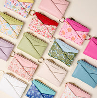 The Darling Effect’s White Luxe Pocket Wallet, crafted from vegan leather, features a gold zipper and key ring. Shown with other compact wallets and cardholders in pastel and floral patterns, neatly arranged on a light background.