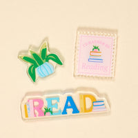 The Darling Effect's All Booked Up Little Magnet Set features three charming acrylic magnets—a plant, a "I'd rather be reading" stamp, and colorful READ letters—perfect fridge decor for any book lover.