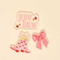The Darling Effect's Howdy Honey Little Magnet Set features three colorful accents: a "yee haw" design in pink with red diamonds, a pair of pink cowboy boots with flowers, and a large pink bow, all on a beige background.