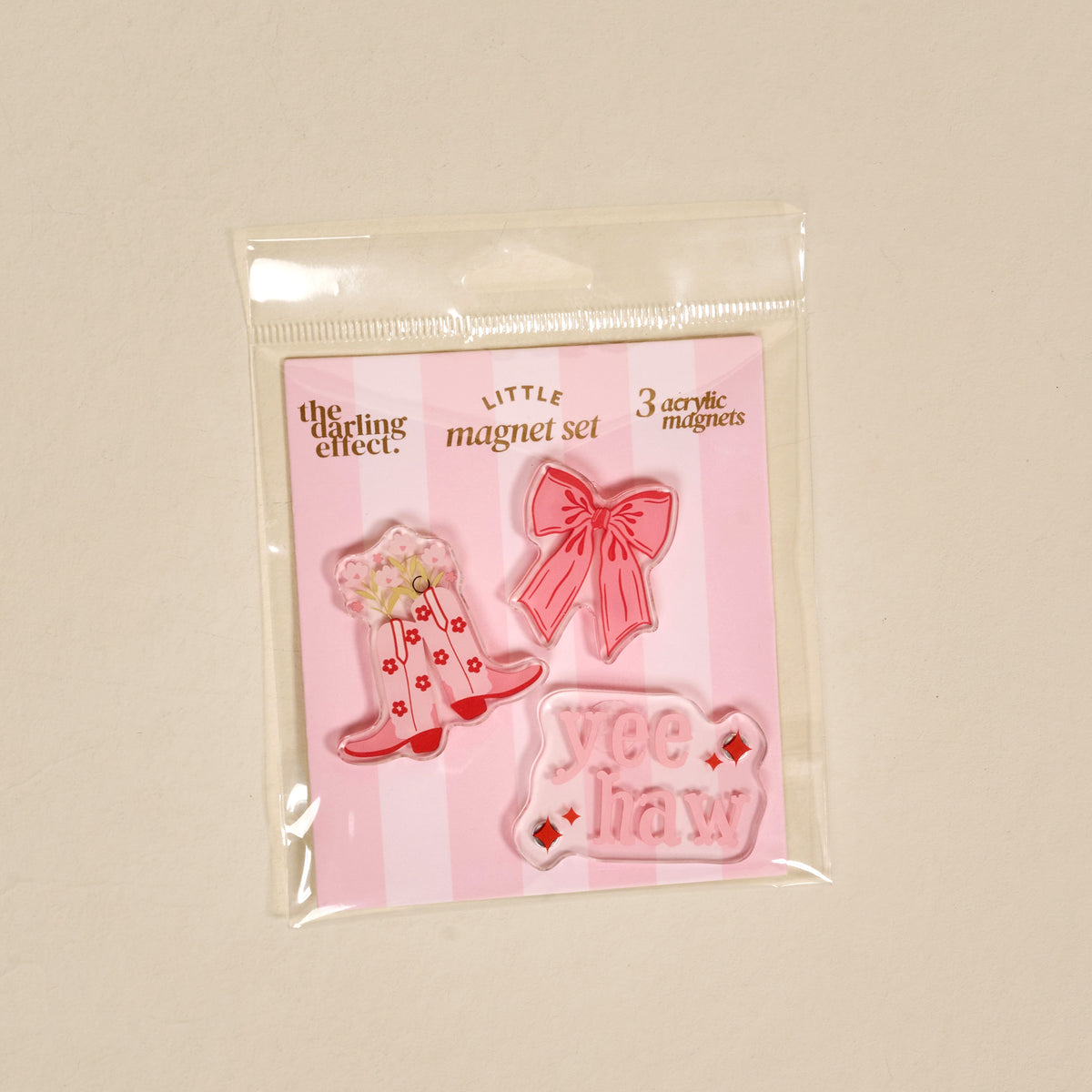 Introducing the Howdy Honey Little Magnet Set by The Darling Effect—a packaged trio of acrylic magnets featuring a cowboy hat adorned with flowers, a pink bow, and a pink “yee haw” sign. Displayed on a cheerful pink-striped backing card, these fridge magnets add a charming decorative accent to any space.