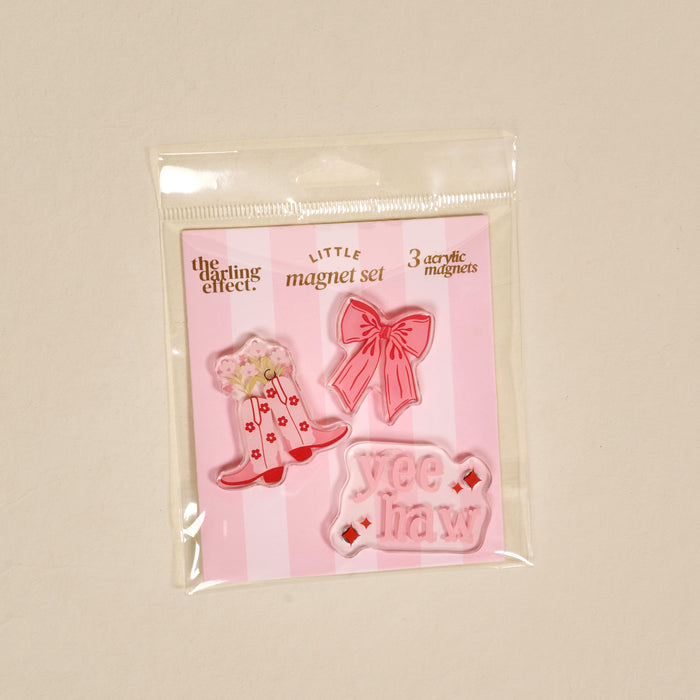 Introducing the Howdy Honey Little Magnet Set by The Darling Effect—a packaged trio of acrylic magnets featuring a cowboy hat adorned with flowers, a pink bow, and a pink “yee haw” sign. Displayed on a cheerful pink-striped backing card, these fridge magnets add a charming decorative accent to any space.