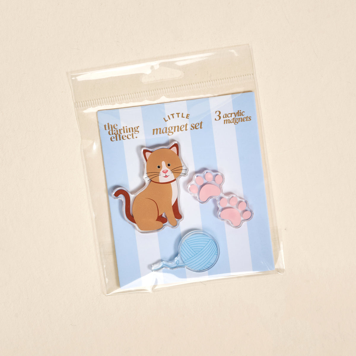 Introducing the Kitty Corner Little Magnet Set by The Darling Effect—a charming package featuring three cute acrylic magnets: a playful cartoon cat, two pink paw prints, and a blue ball of yarn. Perfect for decorating your fridge, this set comes on a pale blue striped background with “LITTLE magnet set” written in cheerful text.