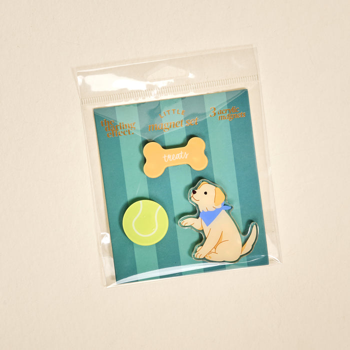 Introducing the Puppy Playtime Little Magnet Set by The Darling Effect: this set includes three playful acrylic fridge magnets packaged in a clear bag—a yellow tennis ball, an orange dog bone labeled “treats,” and a cartoon puppy sporting a blue bandana—each displayed against a green-striped background.