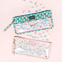 A smiling young woman holds The Darling Effect’s It's Mahj Time Pink Mahjong Tile Keeper Pouch, featuring a clear pocket with assorted Mahjong tiles, against a plain beige background.