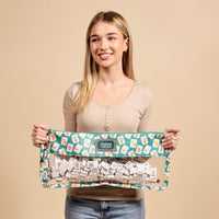 A smiling woman stands against a beige background, holding The Darling Effect's It's Mahj Time Teal Mahjong Tile Keeper Pouch—an eye-catching, patterned pouch ideal for stylish storage of Mahjong tiles.