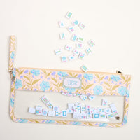 The Darling Effect’s Poppin' Petals Aqua Mahjong Tile Keeper Pouch rests on a cream surface, with Mahjong tiles scattered above and inside its transparent section—ideal for organizing game night essentials.