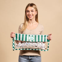 A smiling young woman holds The Darling Effect Sicily Stripes Teal Mahjong Tile Keeper Pouch, featuring green-and-white stripes and a clear front displaying white tiles, against a beige background.