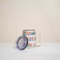 The Darling Effect’s Book Shelfie 14 oz Insulated Mug is a white mug with a large handle, featuring colorful book and plant illustrations. It comes with a clear plastic lid and is shown on a white surface against a neutral background.