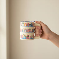 A hand holds The Darling Effect Book Shelfie 14 oz Insulated Mug, featuring colorful book and potted plant illustrations on shelves, set against a plain beige background.