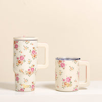 Two cream-colored mugs from The Darling Effect sit side by side on a white surface; one is tall, while the Endless Daydream Cream 14 oz Insulated Mug features double-wall insulation, a clear lid, and pink and green floral patterns.