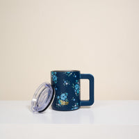 The Darling Effect’s Endless Daydream Navy 14 oz Insulated Mug with a blue floral pattern and square handle sits on a white surface, its transparent lid propped against it. The background is plain and light-colored.