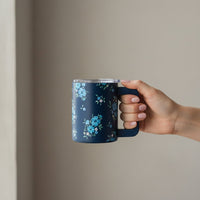 A manicured hand holds The Darling Effect Endless Daydream Navy 14 oz Insulated Mug, featuring a pattern of light blue and white flowers, against a plain, light background.