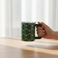 A hand holds The Darling Effect’s Everbloom Forest Green 14 oz Insulated Mug, featuring a blue and white floral pattern, over a light wooden table with sheer white curtains in the background.