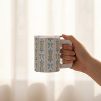 A hand holds The Darling Effect's Oh So Fancy Blue 14 oz Insulated Mug, featuring a blue floral and bow pattern on white, set against a softly lit background with sheer curtains.