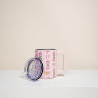 The Darling Effect's Oh So Fancy Pink 14 oz Insulated Mug, with a floral and bow design and pink handle, sits on a white surface. It features double-wall insulation and a clear plastic lid, set against a light beige background.