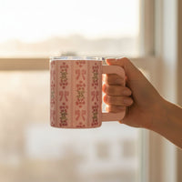 A hand with manicured nails holds The Darling Effect’s Oh So Fancy Pink 14 oz Insulated Mug, featuring floral and bow designs, in front of a sunlit window.
