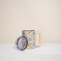 The Darling Effect's Poppin' Petals Aqua 14 oz Insulated Mug, featuring a large square handle and blue floral design with green leaves, sits on a white surface with its clear plastic lid resting beside it against a plain beige background.