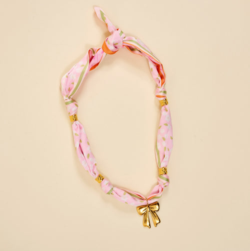 The Darling Effect's Blushing Hour Make a Statement Scarf Necklace features pink floral patterns, gold bow, and beads—an effortlessly bold accessory for any look.