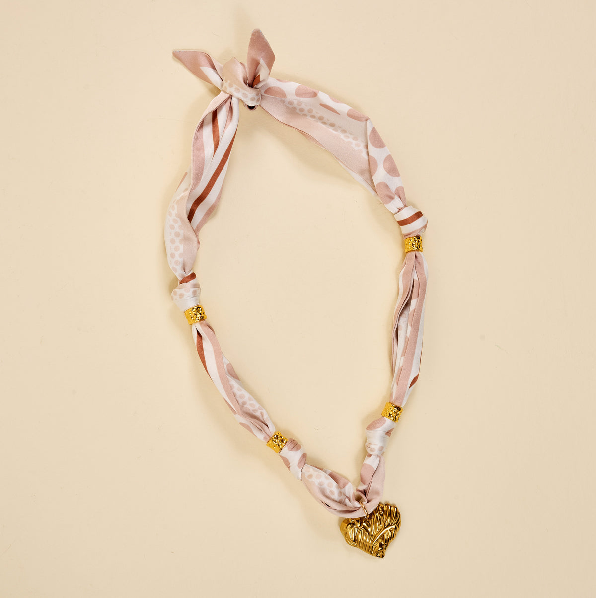 The Darling Effect Desert Dune Make a Statement Scarf Necklace, a pink and white silky accessory with gold bead accents and a heart pendant, is displayed in an oval shape on a beige background.