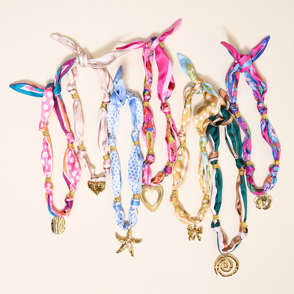 Seven vibrant Desert Dune Make a Statement Scarf Necklaces by The Darling Effect—each crafted from silky fabric and embellished with unique gold charms—are displayed in a row on a light background, creating a striking accessory showcase.