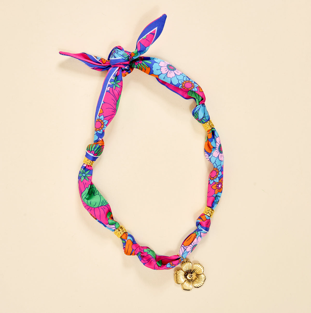 The Darling Effect’s Flower Power Make a Statement Scarf Necklace features artful floral patterns, gold bead accents, knotted details, and a gold flower charm pendant, displayed on a beige background.
