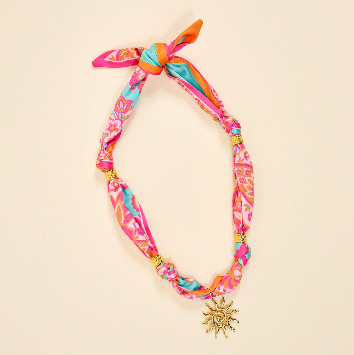 The Darling Effect’s Paisley Glow Make a Statement Scarf Necklace features silky pink, orange, and blue fabric with gold beads, knots, a tie top, and a sparkling gold sun-shaped charm.