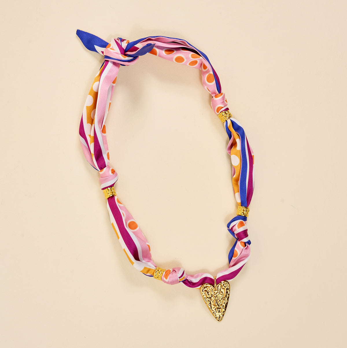 The Darling Effect’s Heart of Gold Make a Statement Scarf Necklace features pink, purple, and orange silk stripes and dots, gold beads, and a textured gold heart pendant for an artful look on beige.