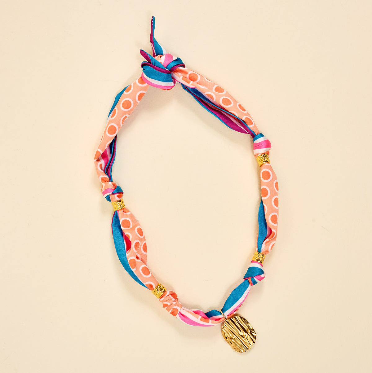 The Darling Effect’s In A Mood Make a Statement Scarf Necklace, in pink, blue, and white with gold beads and a textured gold charm pendant, is displayed on a light beige background.