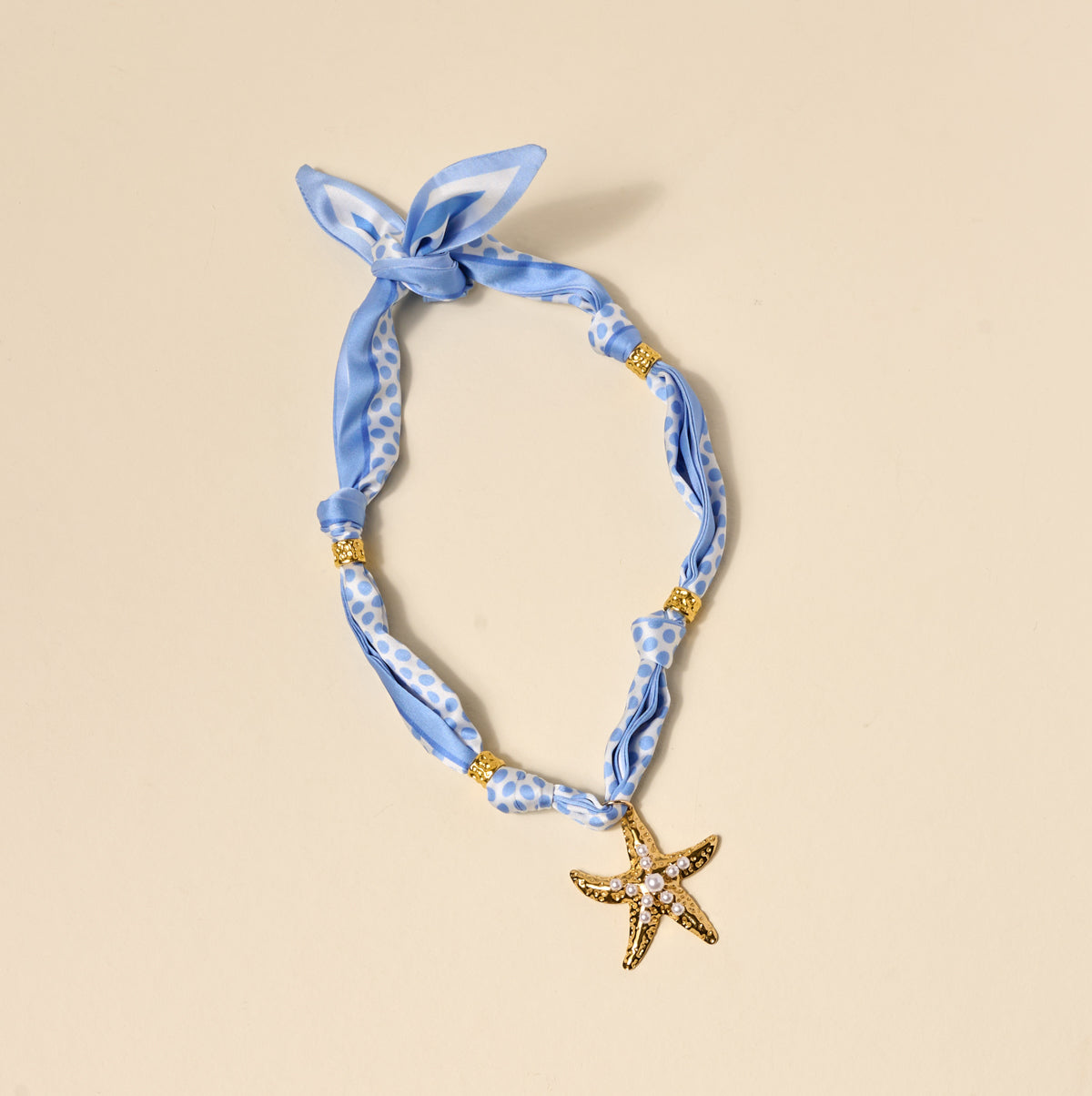 The Darling Effect's Shore Thing Make a Statement Scarf Necklace features artful blue patterns, gold bead details, and a gold starfish charm, shown on a simple beige background.