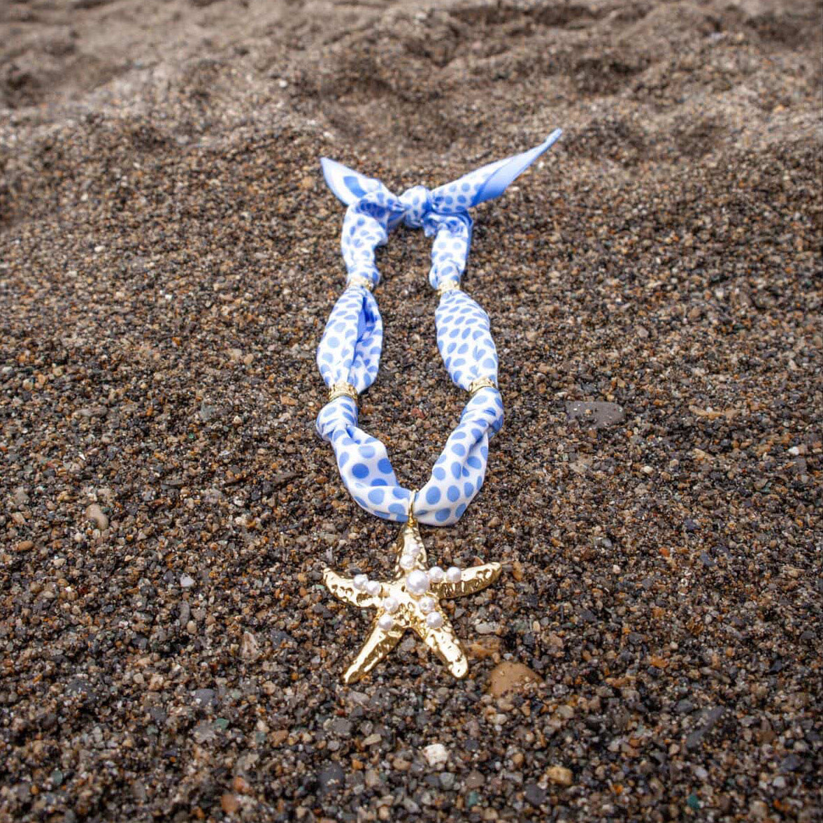 The Darling Effect Shore Thing Make a Statement Scarf Necklace, featuring blue and white polka dots and a gold starfish charm, is displayed on brown sand.
