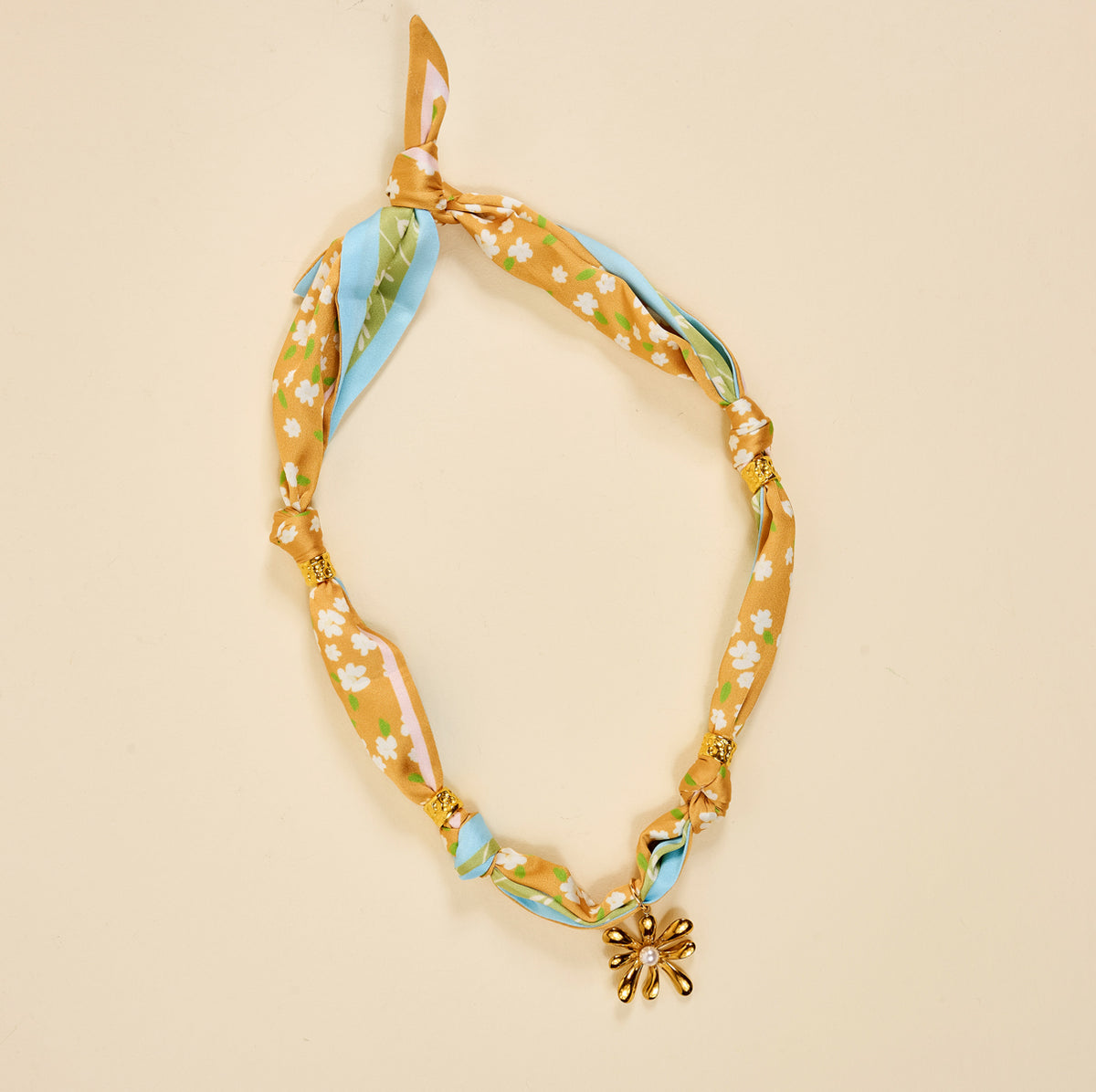 The Sunlit Bloom Make a Statement Scarf Necklace by The Darling Effect features knotted floral fabric in yellow, white, green, and blue with gold beads and a daisy-shaped charm, shown on a beige background.