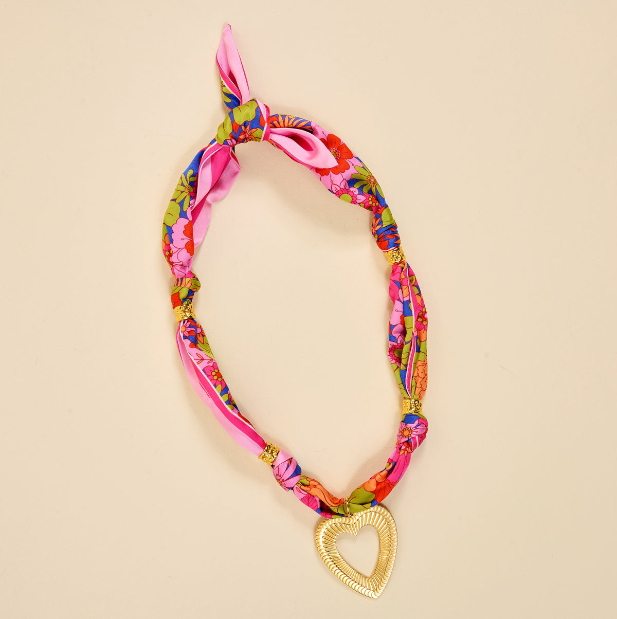 The Darling Effect's Lover's Tempo Make a Statement Scarf Necklace showcases silky pink fabric with colorful florals, finished with a shimmering gold heart pendant—a bold accessory for any look.