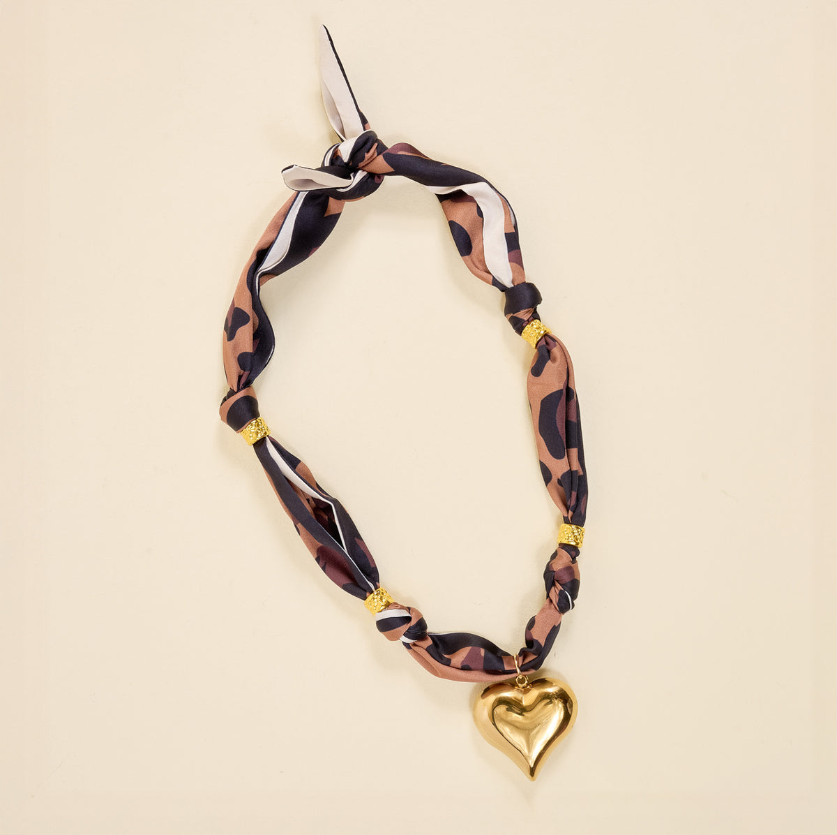 The Darling Effect’s Wild Thing Make a Statement Scarf Necklace showcases artful brown, black, and white patterns with gold accents and a shiny gold heart charm.