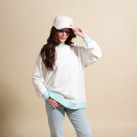 A woman in The Darling Effect’s Bride Oversized Crew and matching baseball cap poses against a beige background, smiling while holding her cap. She has long dark hair and is wearing light blue jeans.