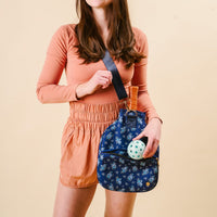 A woman in a peach shirt and shorts carries the-darling-effect's Endless Daydream Navy Pickleball Paddle Case, featuring a floral pattern, racquet slot, space for pickleball paddles and tennis ball, against a beige background.